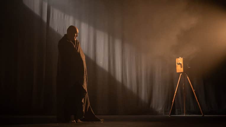 Fishamble presents Silent by Pat Kinevane - a man wrapped in a blanket is looking at old style camera on a tripod