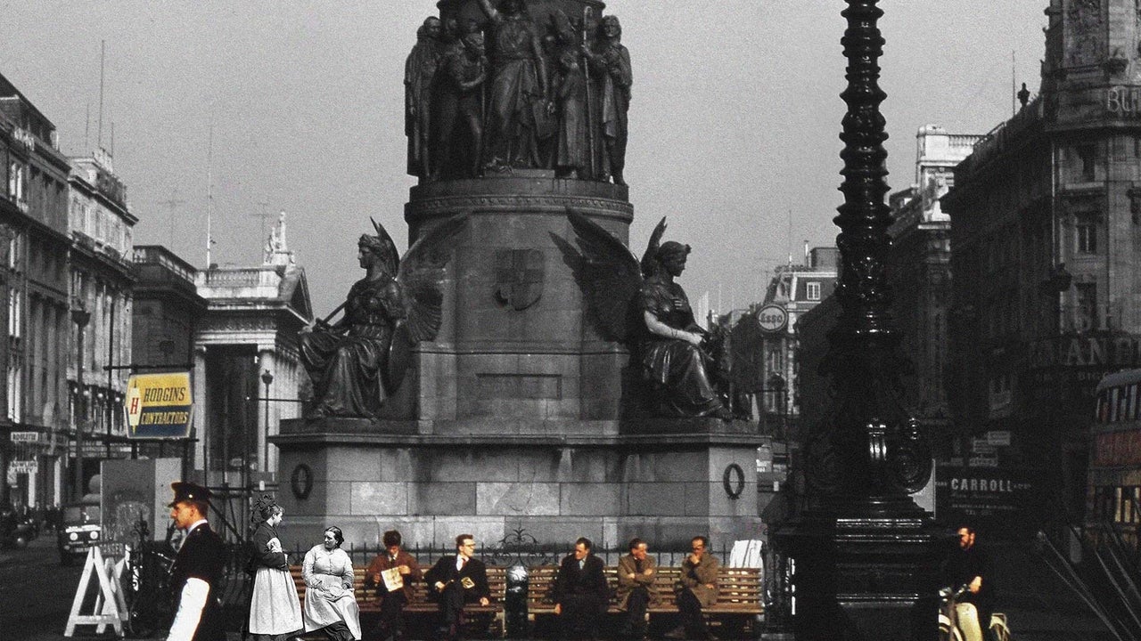 People scattered along 2 benches underneath plinth of large monument in a city in the past.