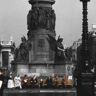 People scattered along 2 benches underneath plinth of large monument in a city in the past.