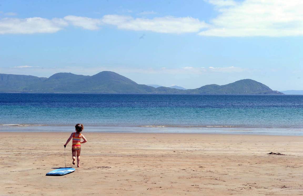 A little girl running on a sandy beach towards the water with the mountains in the distance and the blue sky above her.