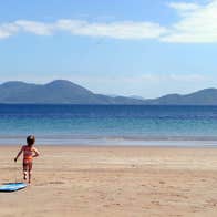 A little girl running on a sandy beach towards the water with the mountains in the distance and the blue sky above her.