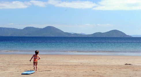 A little girl running on a sandy beach towards the water with the mountains in the distance and the blue sky above her.