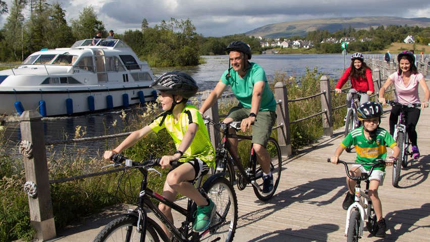 Two adults and three children on bikes on a boardwalk adjacent to a river with a boat on it