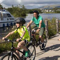 Two adults and three children on bikes on a boardwalk adjacent to a river with a boat on it