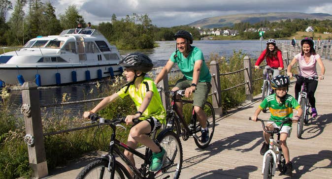 Two adults and three children on bikes on a boardwalk adjacent to a river with a boat on it