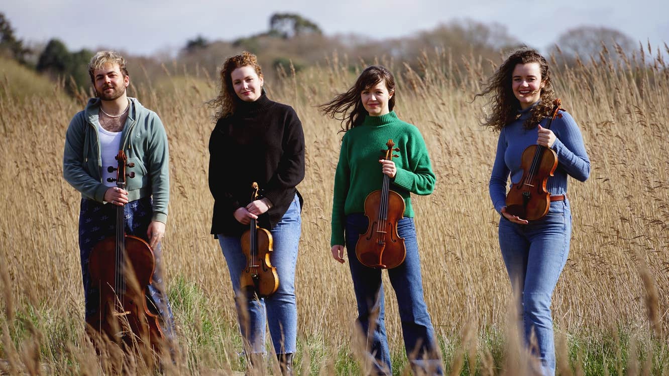 The members of the Calathea Quartet holding their instruments. They're standing on a path surrounded by tall grass.
