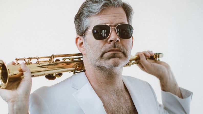 A man in sunglasses and a white open jacket showing a hairy chest is holding a gold coloured wind musical instrument over his shoulders.