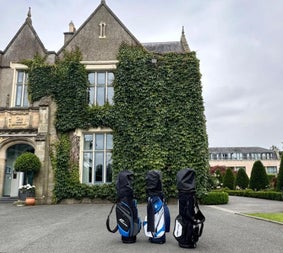 Three golf club bags standing in front of a large house with windows and ivy climbing up one side