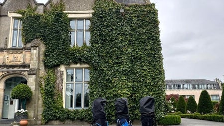 Three golf club bags standing in front of a large house with windows and ivy climbing up one side