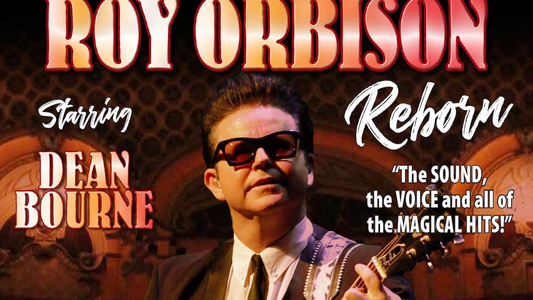 A man wearing dark sunglasses in a suit is playing a guitar.