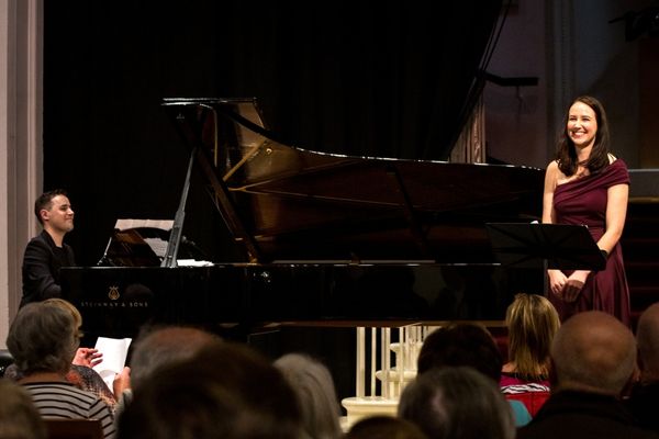 A man sat at a grand piano with a woman in evening dress at the end of the piano smiling facing the audience