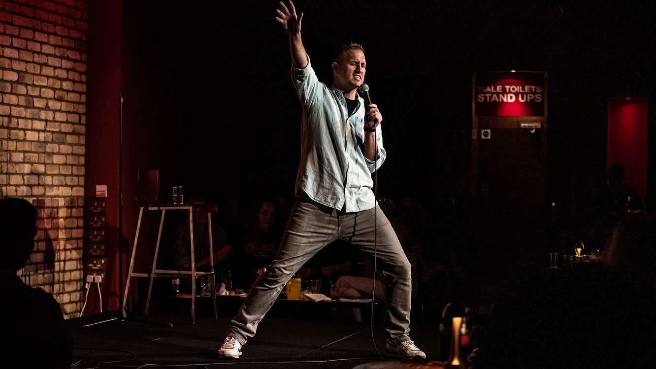 A comedian on stage holding a microphone with his right arm and leg extended