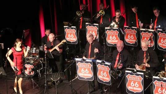 Photo of band members in 2 rows wearing black shirts and red ties sitting or standing in front of music stands with the red route 66 logo on, to left of stage a woman in a red dress holding a mic is dancing.