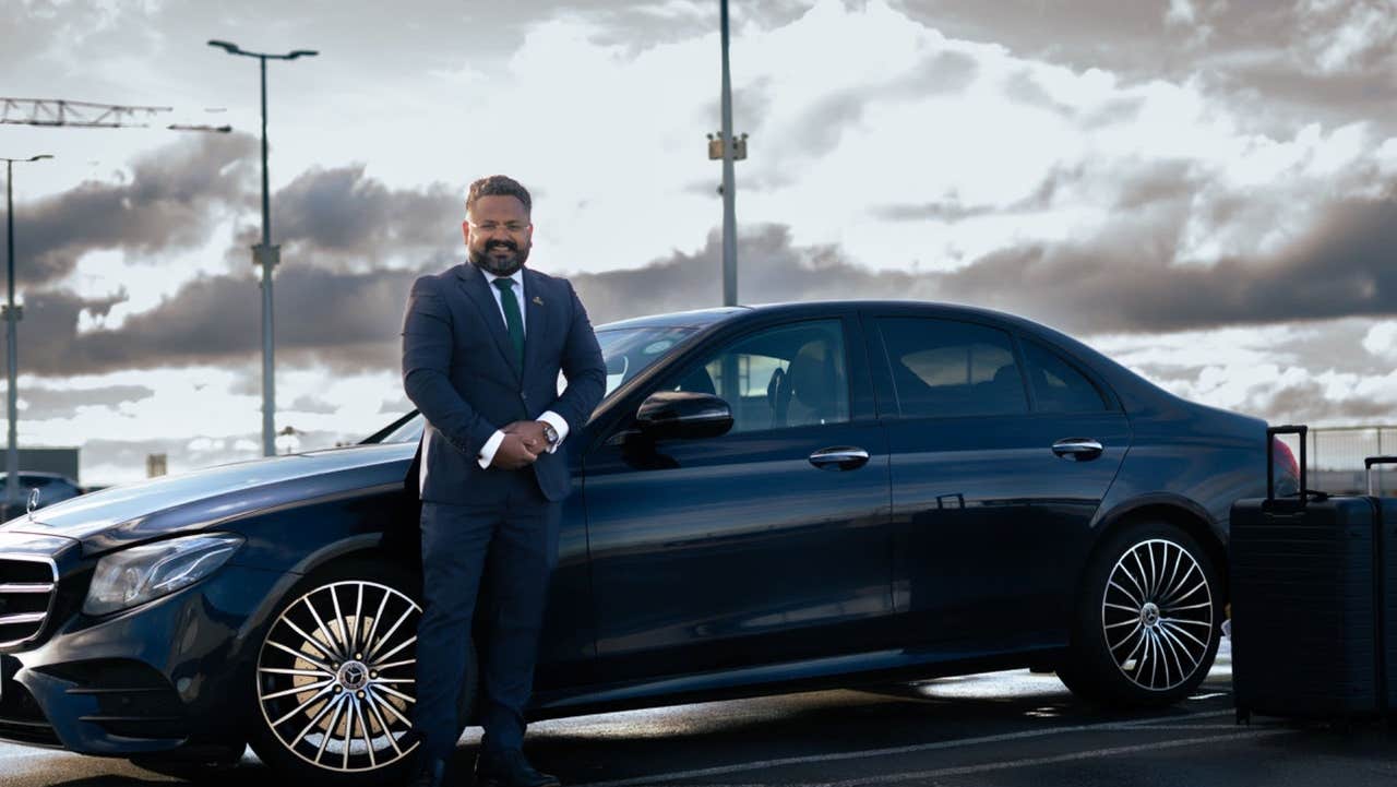 A driver stands beside a dark car in a parking area under a cloudy sky