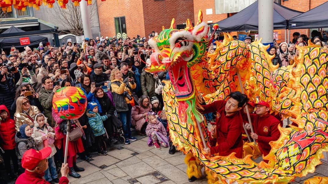 Large, yellow, red and black dragon operated by 2 men in red with large crowd watching on.