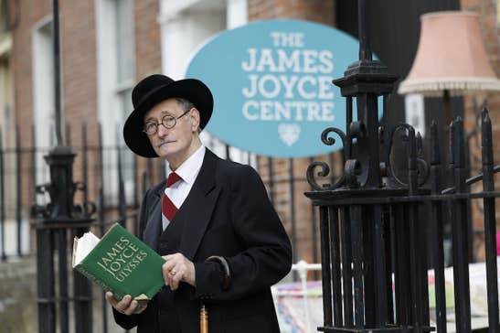 A man in black suit and hat is holding a green book, with black ornate railings and georgian brick building behind him.