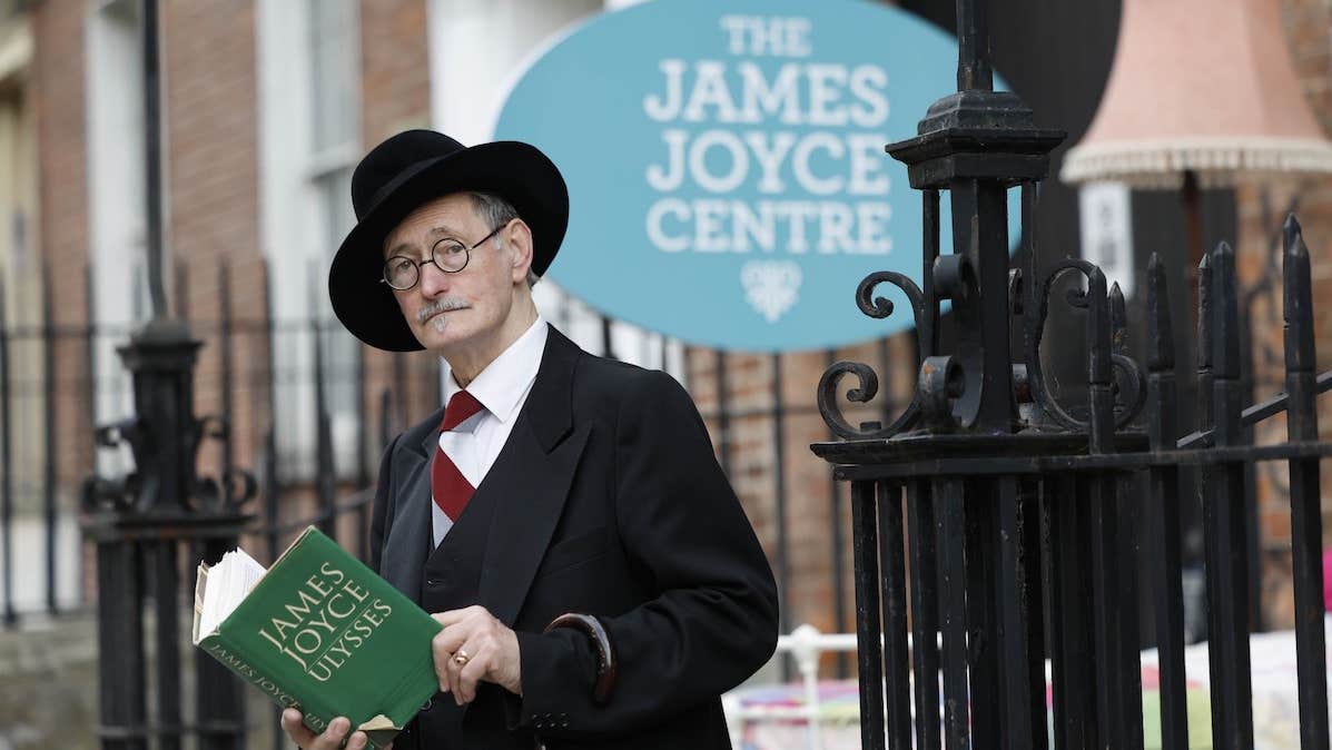 A man in black suit and hat is holding a green book, with black ornate railings and georgian brick building behind him.