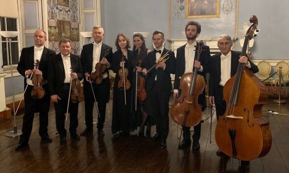 A group of 8 musicians in black evening dress with stringed instruments standing in a line in an old room.