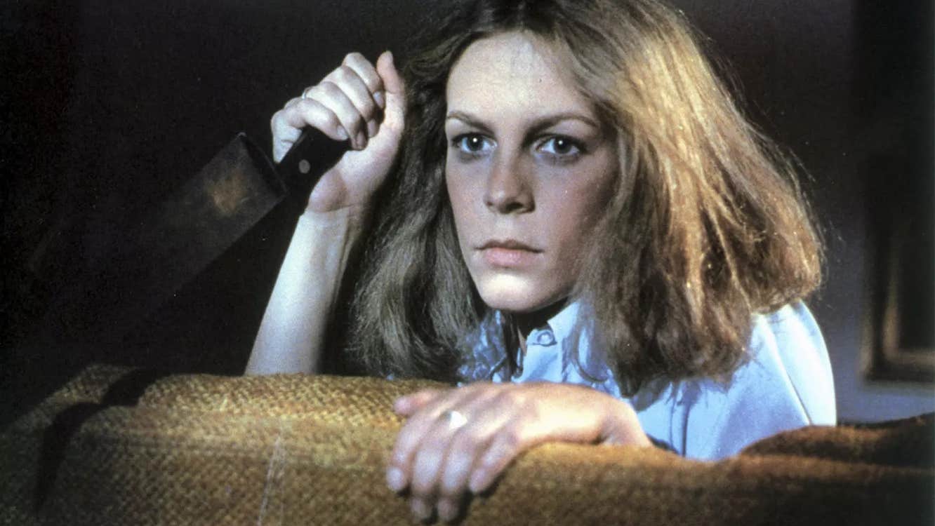 A woman with a kitchen knife is hiding behind a piece of furniture.