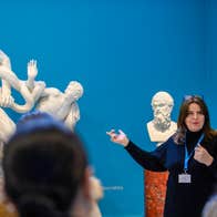 A tour guide is pointing towards a white sculpture of classical origin with blurred backs of heads in the foreground, against bright blue wall in background.