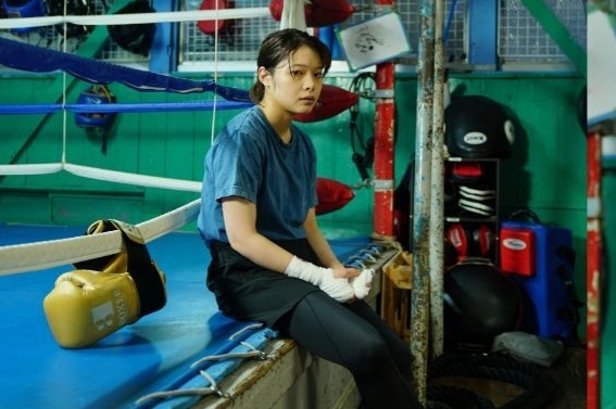 A young woman is sat on the edge of a boxing ring looking worried.