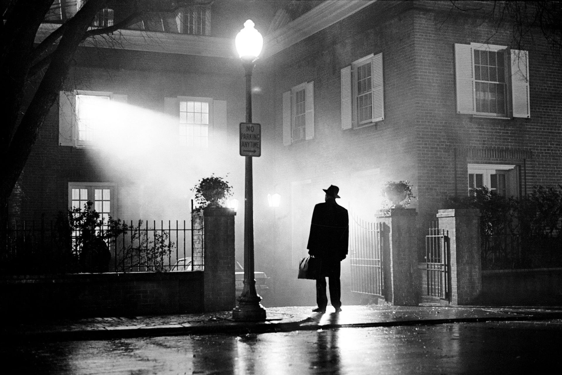 A black-and-white image of a man standing in front of a building. A bright light is shining from one of the windows at the man, making him a silhouette. It has a spooky atmosphere.