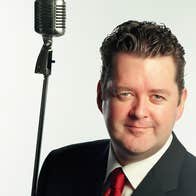 A man is smiling, in a dark jacket with red tie, beside is old fashioned style mic on a tall stand, against plain white background.