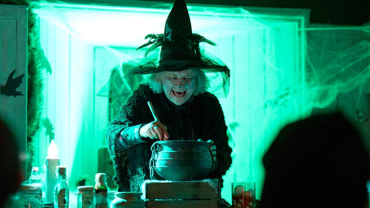A person wearing black with tall, pointy hat is stirring a small black pot on a steaming block with small bottles, all lit from behind with bright green light.