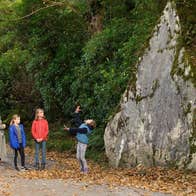 One adult and three children standing amongst fallen leaves with a rock and trees behind them