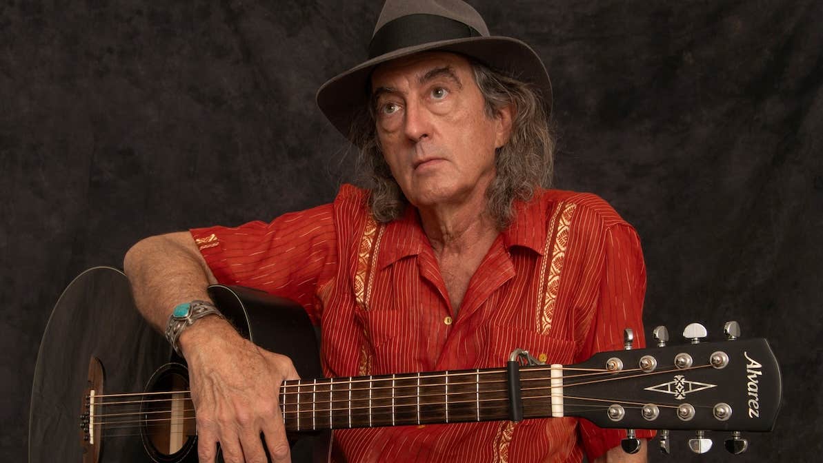 A man in red shirt and grey hat is holding a guitar.