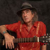 A man in red shirt and grey hat is holding a guitar.