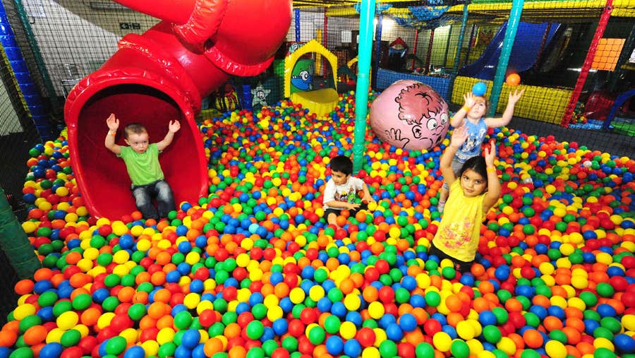 Three children standing in soft ball pit with another child just coming out of a red slide just about to land into the soft ball pit