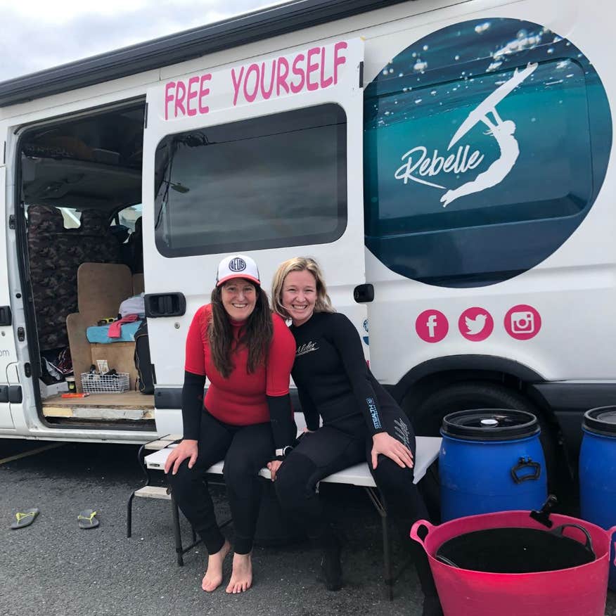 Two women smiling in surfing gear in front of a white van with Rebelle Surf logo in Sligo.