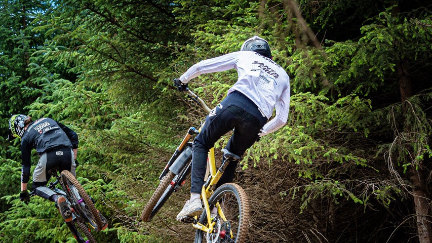 Two bikers mid jump on a forest track beside trees at Glencullen Adventure Park