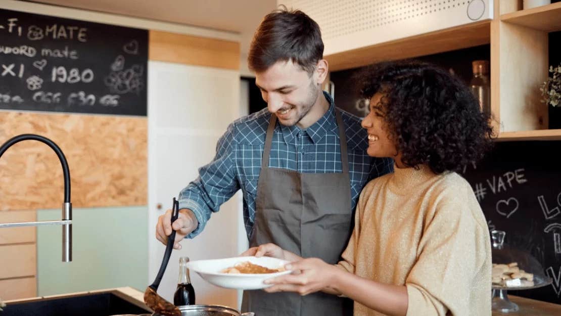 A couple are smiling, standing in a kitchen cooking.