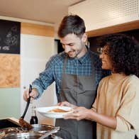 A couple are smiling, standing in a kitchen cooking.