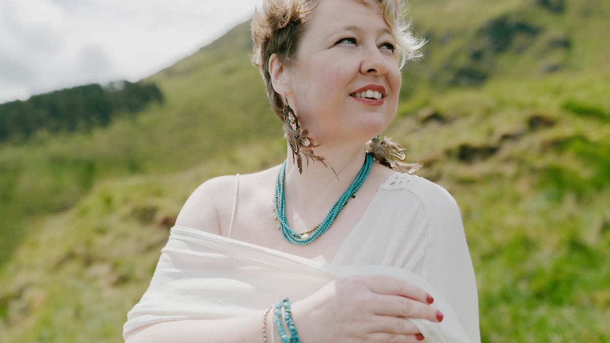 Photo: Berit Alits. A woman is wrapping a white shawl around her shoulders looking away to her left, outdoors with grassy hill in the background.
