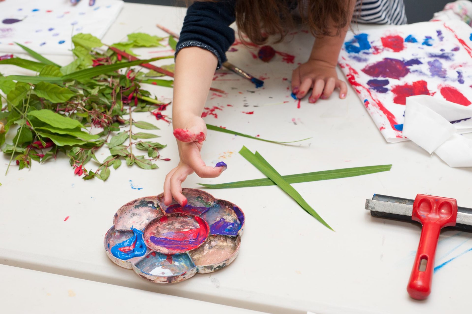On a white table top, a child's hand and arm are reaching across into a plate of paints with plant leaves and paper beside.