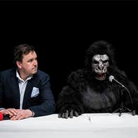 A man in a suit and a person in a gorilla suit sitting beside each other at a table with a black background