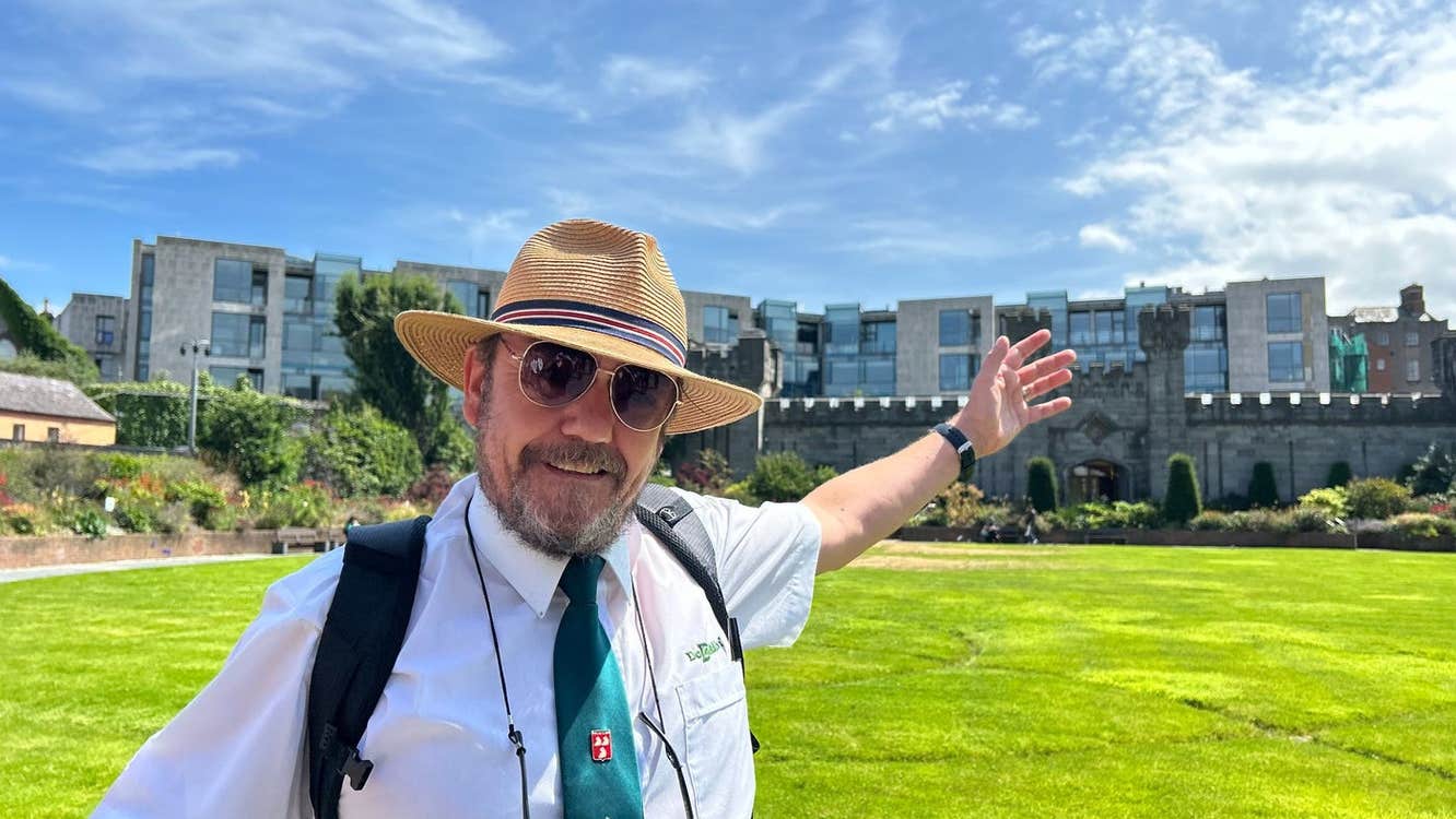 A tour guide looking at the camera while pointing at buildings to his left