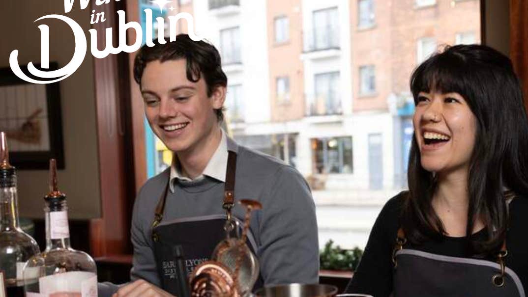 2 people are at a bar counter laughing with cocktail equipment in front of them.