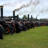 Long line of black steam engines into the distance, smoke coming out of the chimneys, small groups of people in front of each engine, all in a grassy field.