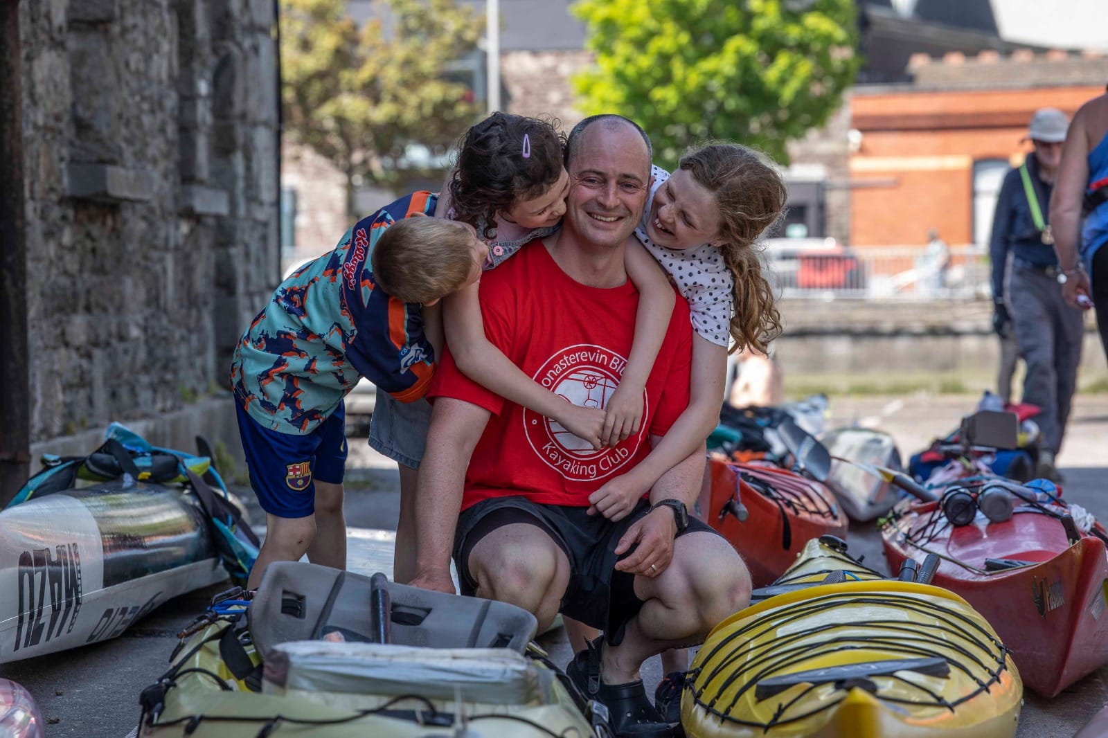 An emotional man in red T-shirt is crouched down surrounded by canoes with 3 children all hugging him and smiling.