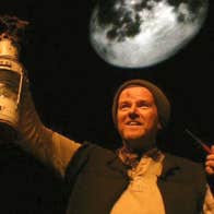 A black and yellow photo against dark background of a man in rough clothes and wooly hat holding a pipe in one hand, an old oil lamp up in the other.