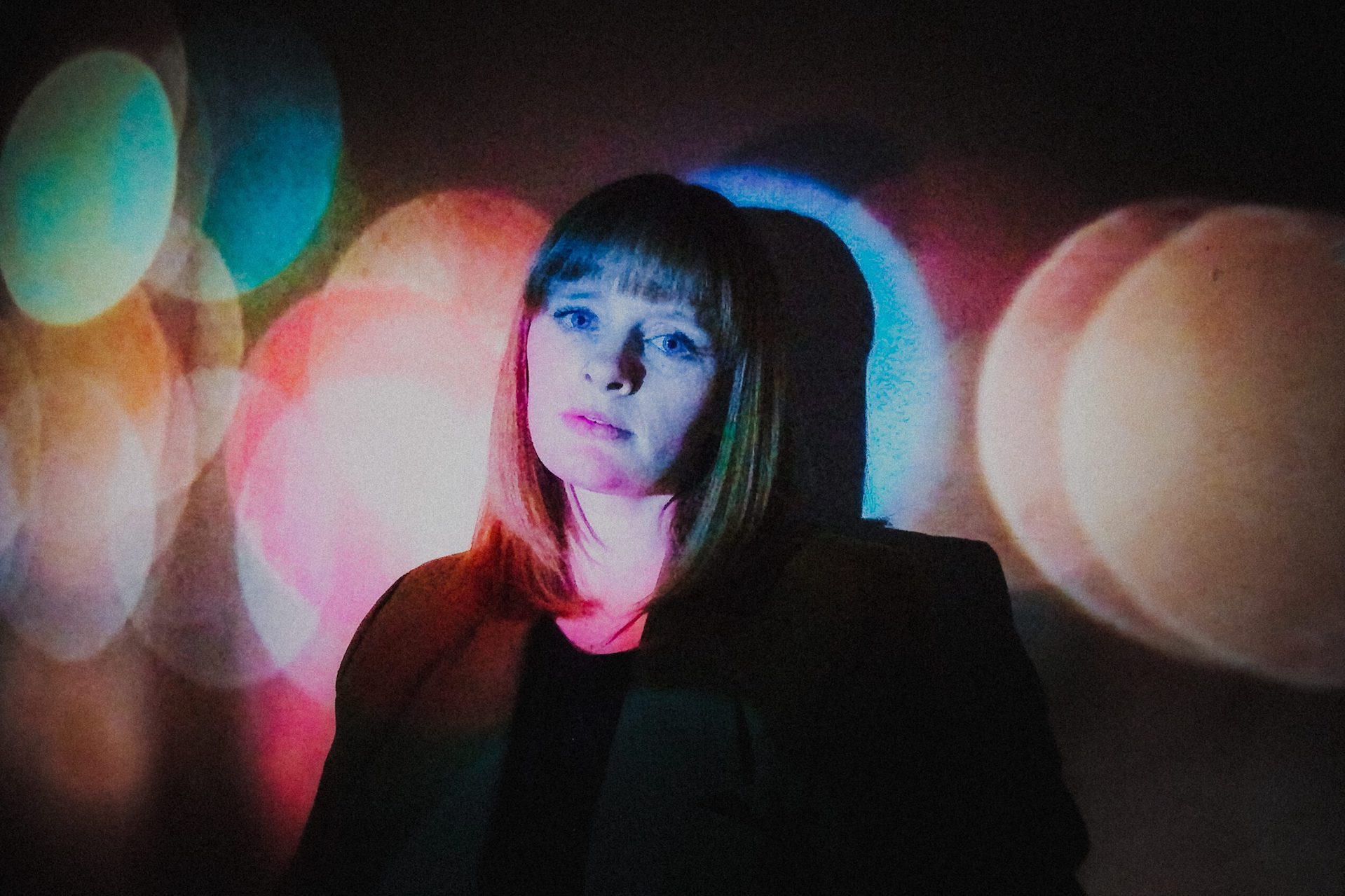 A woman is looking slightly sadly at the camera with faded colourful spot lights on and around her.