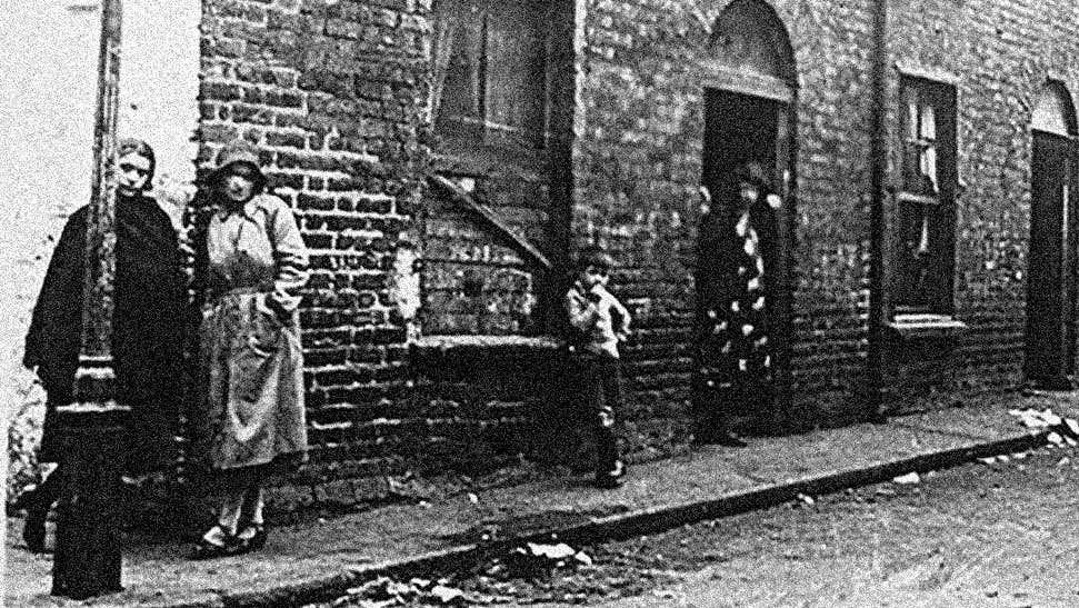 Very old black and white photo of 2 women standing at corner of row of old, brick terraced houses.