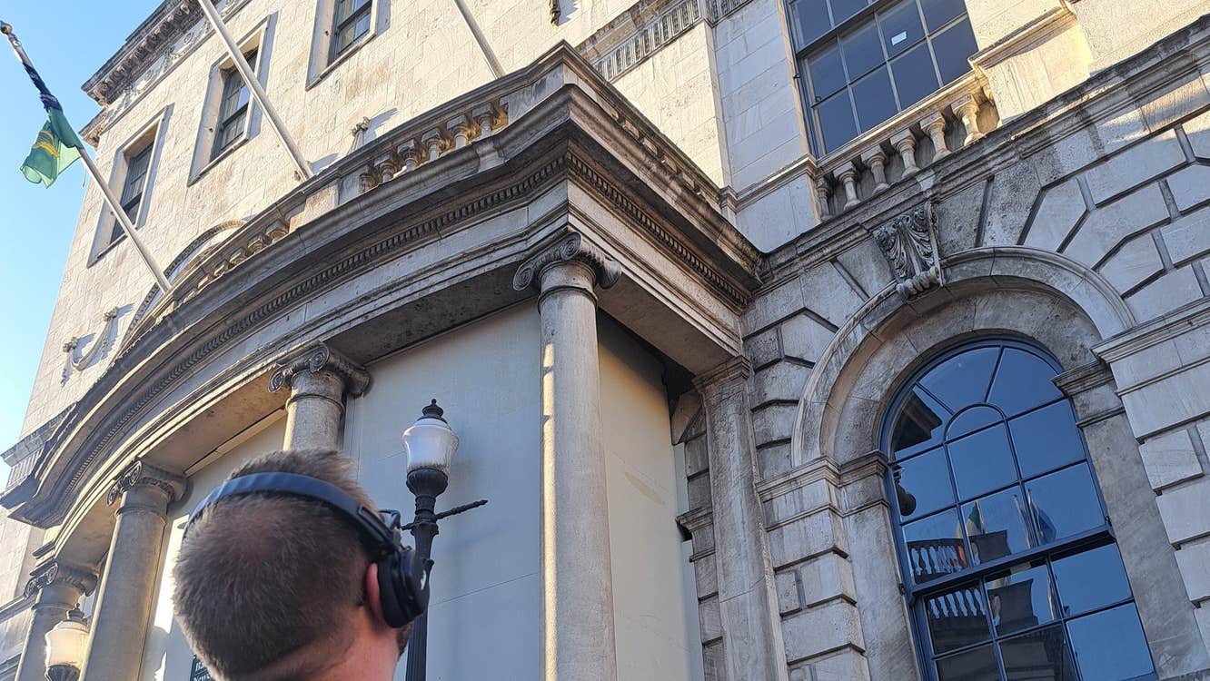 Person wearing headphones holding a phone looking up at a building with three flags flying