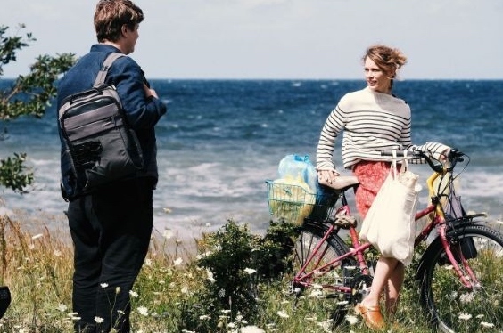 A man with a rucksack is talking to a woman on a bike with shopping in a grassy area with the sea in the background.