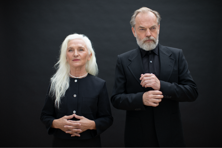 Starring Hugo Weaving and Olwen Fouéré. A woman dressed in black with white buttons and necklace and white hair is standing beside a man dressed in black shirt and jacket looking serious, against plain dark background.