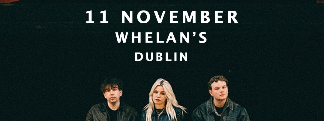 3 people dressed in black clothes and jackets are seated on the edge of a wooden stage.